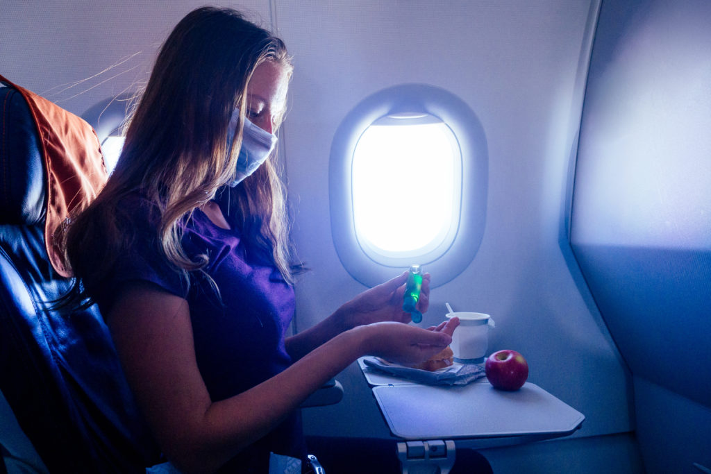 young woman sitting in airplane window seat putting on hand sanitizer while wearing a face mask